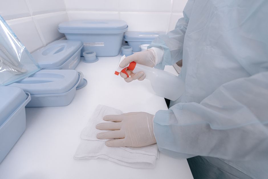 A healthcare worker wearing gloves sanitizes a surface with a spray bottle in a medical setting.